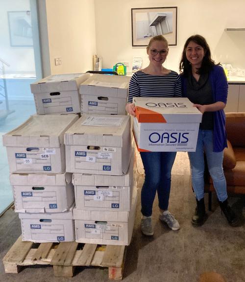 The OED manager and officer holding an archive box while standing close to a pile of 14 other archive boxes}
